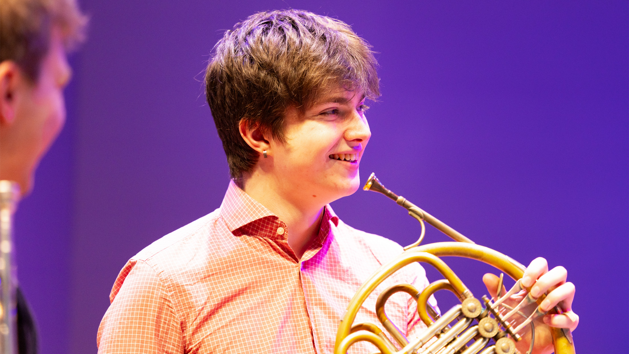 A male student holding a French horn, smiling in a group. A male student holding a French horn, smiling in a group.